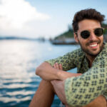Handsome smiling man wearing sunglasses and looking away. He is sitting by the sea